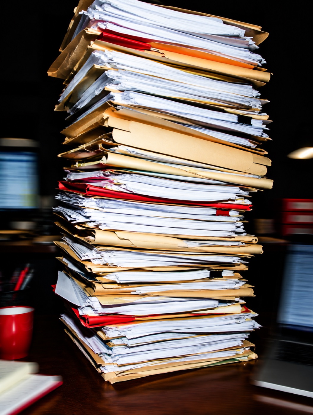 A leaning tower of printed paperwork and manila folders on an office desk, lit by a hard flash.